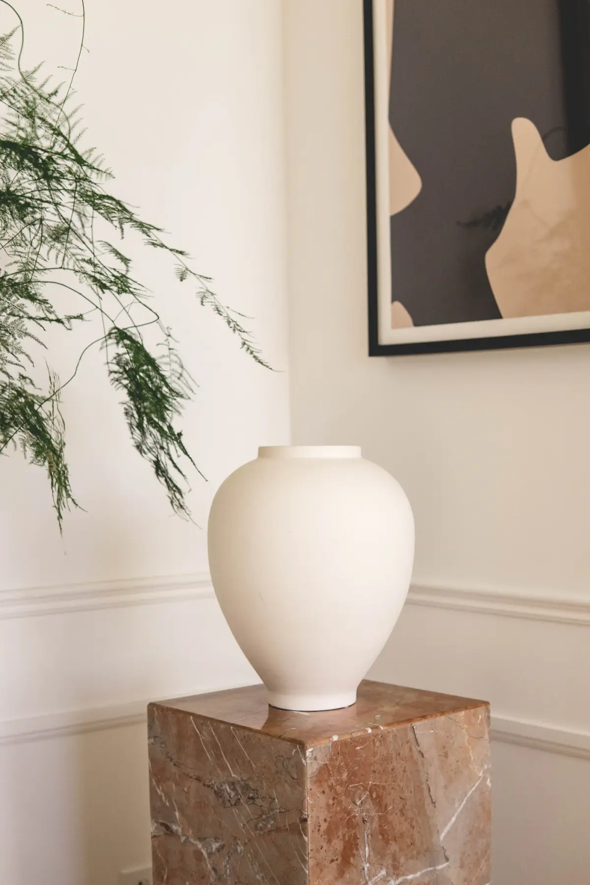 A tall round-shouldered chalk-white vase on a wooden stool beside a trailing plant