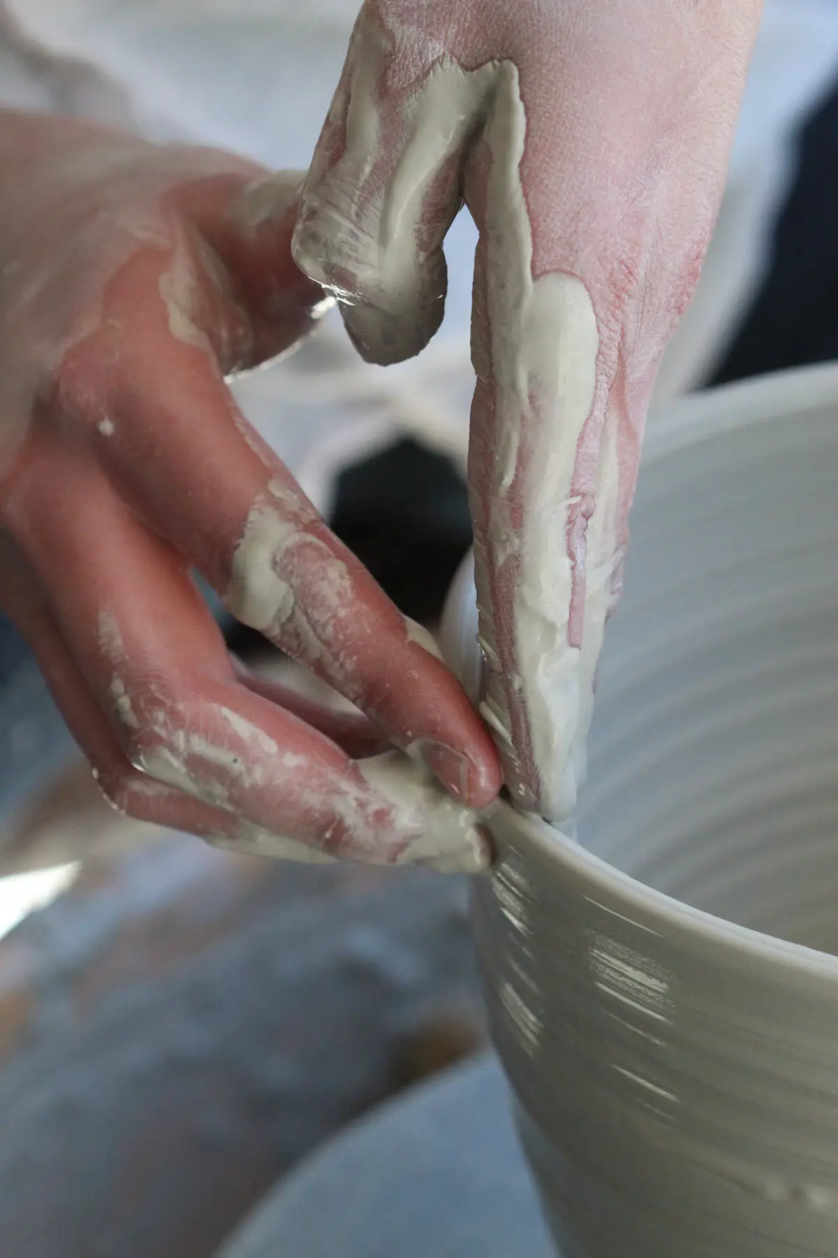 Hands covered in wet grey clay pulling up the wall of a pot