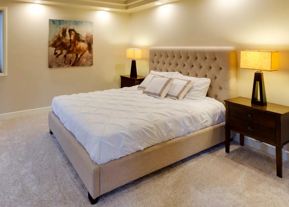 The Haven bed in sand fabric with a tall buttoned headboard and white bedding, beside two bedside lamps