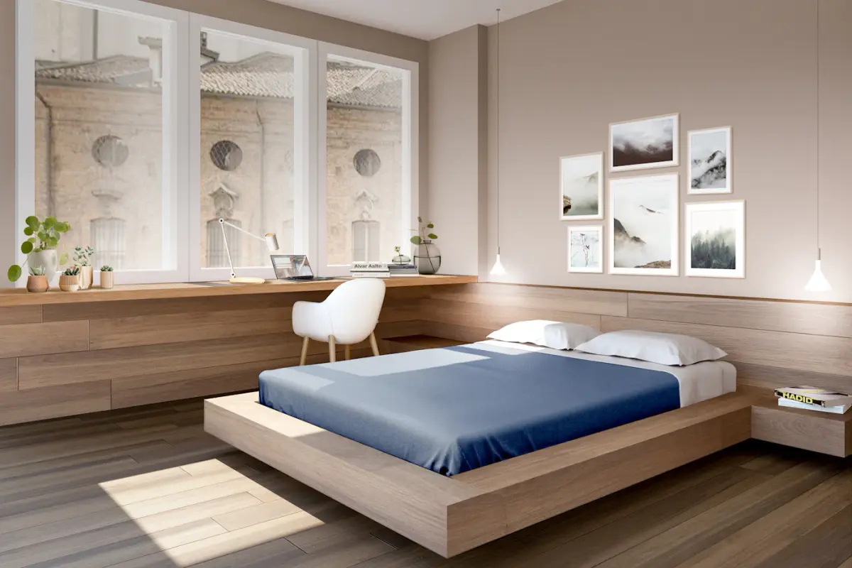 A low oak platform bed with a navy blue cover in a bright room with framed prints and a desk