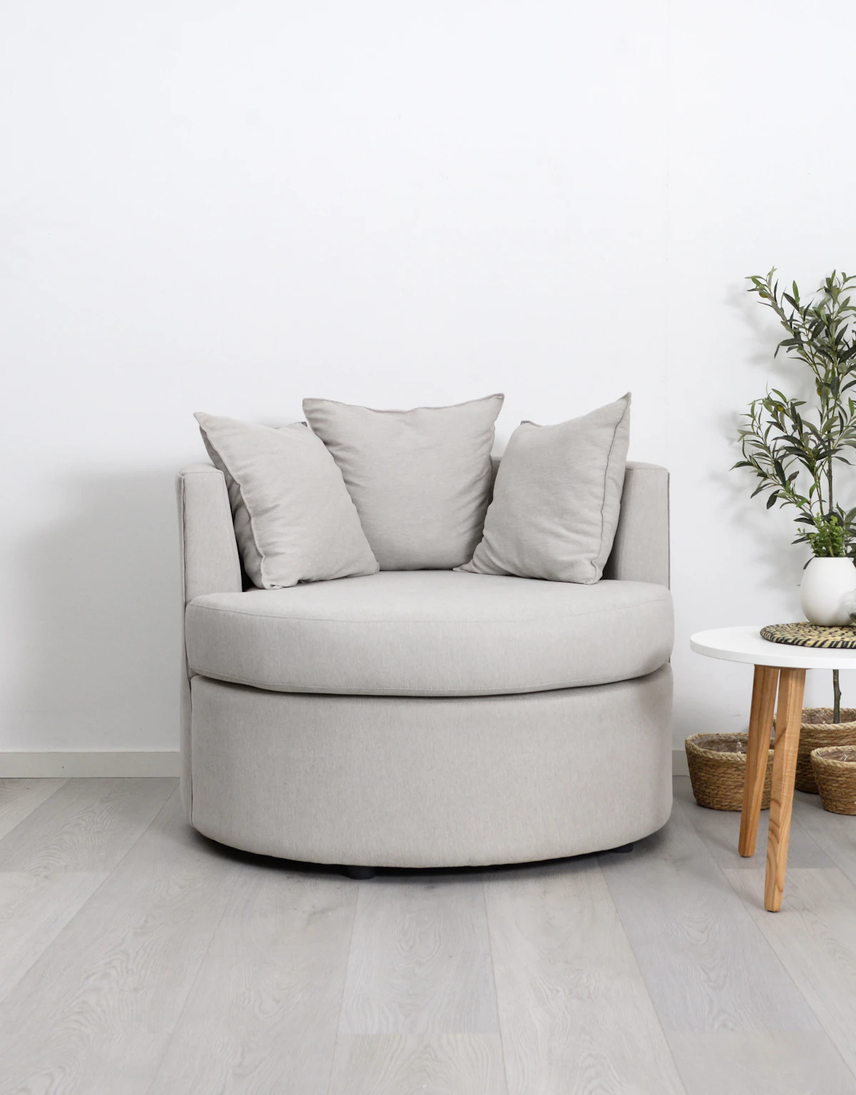 A round light grey armchair with two loose cushions beside a small wooden side table