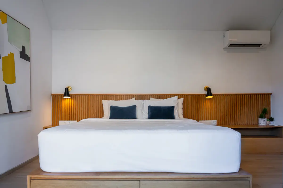 A bed with a white mattress and navy cushions against a slatted wooden headboard wall with two wall lamps