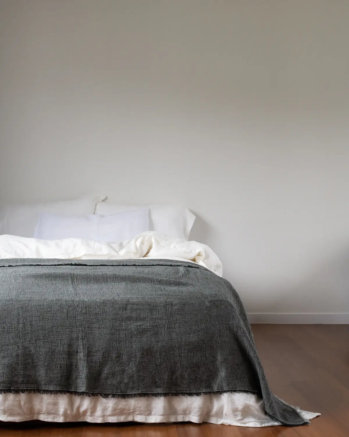 A neatly made bed with white pillows and a grey knitted blanket against a plain white wall