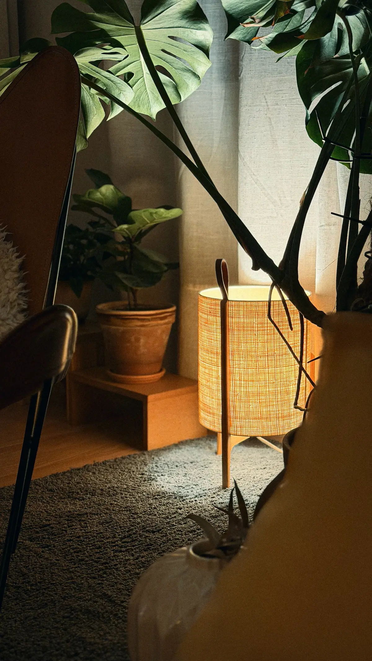 A glowing woven linen column lamp on slim legs beside potted plants in a warm corner