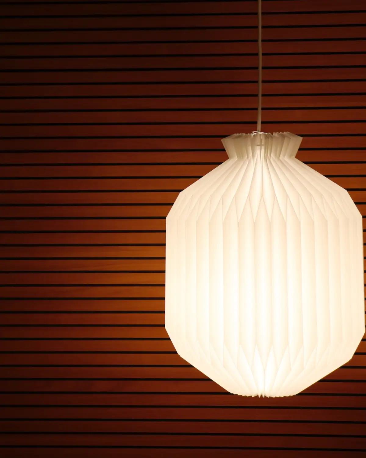 A lit white pleated paper pendant against a wall of dark wooden slats