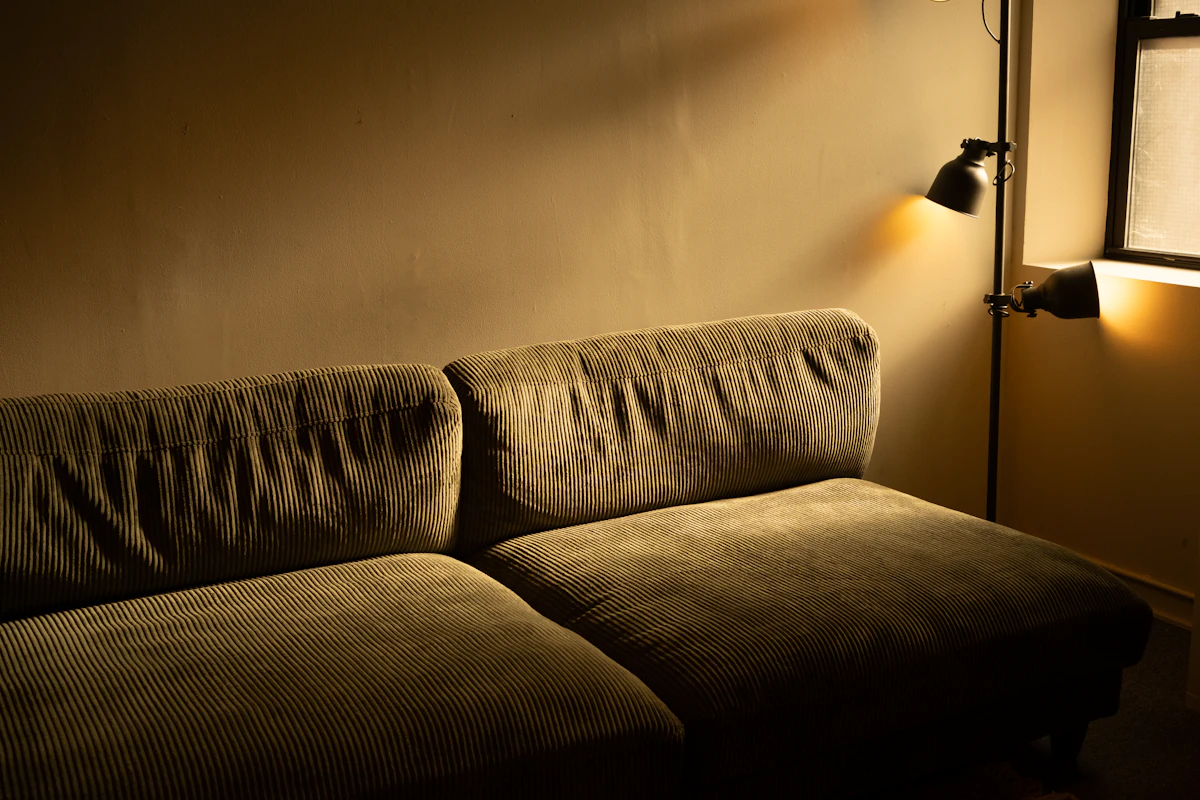 A corduroy sofa in warm evening light, lit by a two-headed black floor lamp behind it